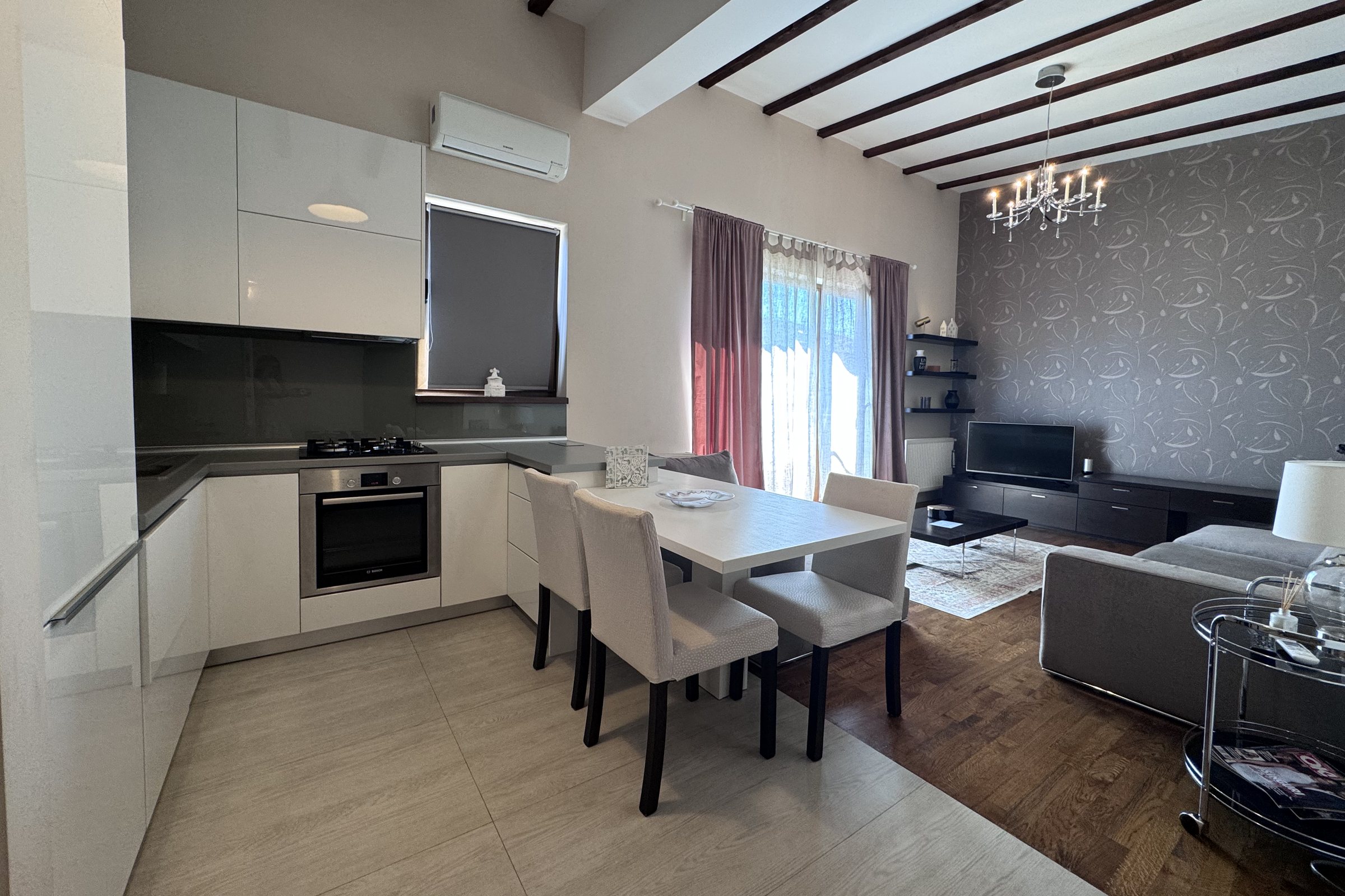 Zona de zi a apartamentului de pe Eugen Ionesco: bucătăria, masa și livingul într-un singur spațiu, sub tavanul cu grinzi de lemn